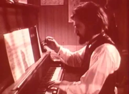 The still from *Erik Satie and the King of the Beans - Part I* features a man seated at a piano, visibly engaged in playing music or composing. He has a beard and is dressed in period-appropriate attire, including a vest. The setting appears to be a cozy room, adorned with decorative elements on the walls. The scene captures a moment of artistic focus, reflecting Satie's creative process in a warm, sepia-toned aesthetic typical of historical educational films.