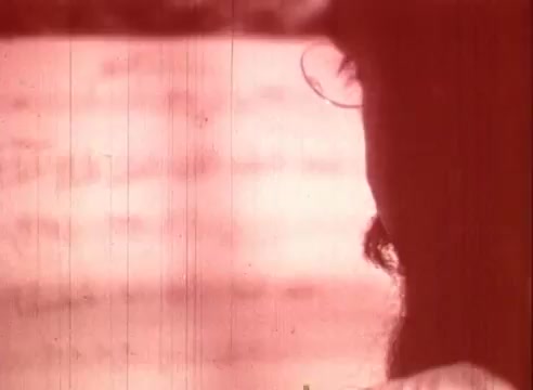 The still from 'Erik Satie and the King of the Beans - Part I' features a close-up profile of a figure, likely depicting Erik Satie, set against a blurred, warm-toned background. The film grain and reddish tint add a vintage quality, enhancing the educational context of the piece. The focus on the figure suggests introspection, aligning with Satie's artistic themes.