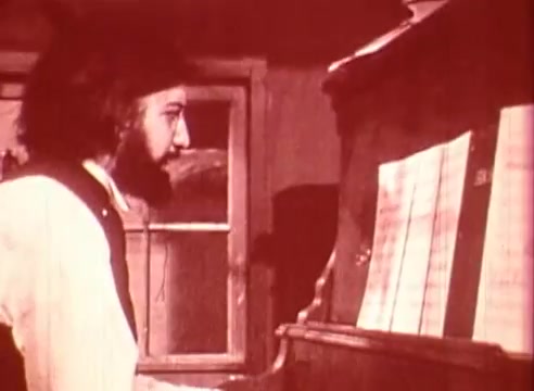 The still captures a character, likely portraying Erik Satie, seated at a piano. The lighting has a warm, vintage tone, enhancing the educational ambience of the film. The character's expression seems focused as he looks at sheet music, suggesting a moment of creativity or contemplation. The background features a window, hinting at a domestic setting, indicative of Satie's personal life and artistic environment.