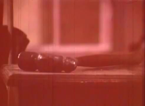 The still from 'Erik Satie and the King of the Beans - Part I' features a close-up of a wooden surface with a couple of objects on it. One object appears to be a rounded, possibly bean-shaped item, while another, longer object is also in view, hinting at an artistic or whimsical context. The color tone is reddish, contributing to a vintage or nostalgic feel typical of 16mm film. The composition suggests an emphasis on texture and form, aligning with the film's educational purpose related to Satie's artistic themes.