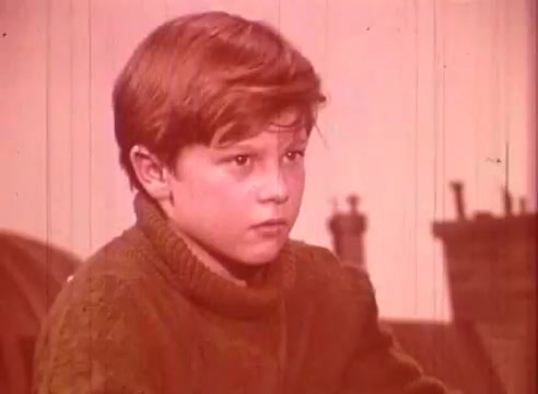 The still from 'Erik Satie and the King of the Beans - Part I' features a young boy with a serious expression, wearing a knitted sweater. The image has a reddish hue, creating a nostalgic atmosphere. In the background, there are rooftops, hinting at an urban setting. The focus is on the boy's thoughtful demeanor, suggesting a moment of contemplation or introspection.