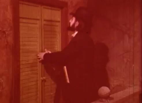 The still from 'Erik Satie and the King of the Beans - Part I' features a man dressed in period attire, including a bowler hat. He is standing near a closed set of wooden shutters painted in a vibrant yellow. The lighting casts a warm, reddish hue over the scene, adding an atmospheric quality. The man's posture suggests he is about to open the shutters, hinting at a moment of anticipation or discovery within the narrative of the educational film.