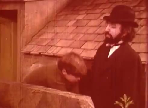 The still from 'Erik Satie and the King of the Beans - Part I' depicts a scene featuring two characters, one dressed in a vintage black coat and top hat, and the other, possibly a younger figure, bending toward a low wooden structure, suggesting a moment of interaction or inquiry. The sepia-toned color palette conveys a historical or whimsical aesthetic, typical of educational films exploring artistic themes. The setting appears rustic, with a roof made of shingles in the background, enhancing the film's vintage feel.