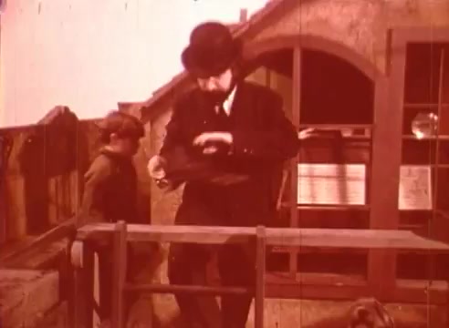 The still from 'Erik Satie and the King of the Beans - Part I' depicts a man in a top hat and formal attire interacting with a young boy, who appears curious or attentive. The background features a rustic setting, likely suggesting a whimsical or educational theme related to Satie. The color tones are warm and slightly faded, reminiscent of vintage film techniques. The scene captures a moment of engagement between the two characters, set against a charming, storybook-like environment.