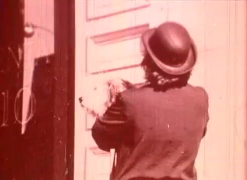 The still from 'Erik Satie and the King of the Beans - Part I' depicts a person wearing a bowler hat holding a small dog while standing in front of a door. The scene likely conveys a whimsical or humorous interaction and is shot in a vintage style characteristic of 16mm films, contributing to its educational and artistic atmosphere. The color palette appears muted, enhancing the nostalgic feel of the film.