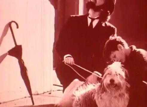 The still from 'Erik Satie and the King of the Beans - Part I' features a man in period clothing and a hat, appearing contemplative, alongside a boy who seems relaxed, possibly leaning against him. A dog sits nearby, adding a whimsical element to the scene. An umbrella lies on the ground, suggesting a casual outdoor setting. The overall tone is light and nostalgic, reflective of the film's educational focus on Satie's music.