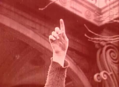 The still features a hand raised with one finger pointing upward, set against a backdrop that suggests an architectural structure. The film has a reddish tint, contributing to its vintage aesthetic. This moment likely emphasizes a significant theme or concept within the educational narrative of 'Erik Satie and the King of the Beans - Part I.'