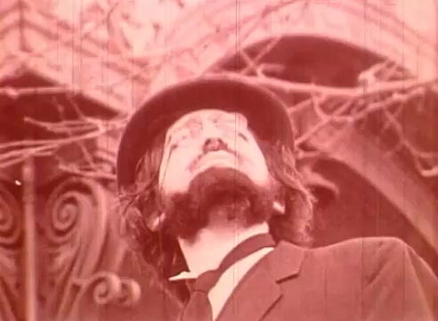 The still from 'Erik Satie and the King of the Beans - Part I' features a man with a prominent beard and mustache, wearing a bowler hat and a suit. He gazes upward with an expression of contemplation or inspiration. The background suggests a historic or artistic setting, possibly featuring architectural elements and vines. The film's aesthetic appears vintage, contributing to its educational and cultural theme centered on the composer Erik Satie.