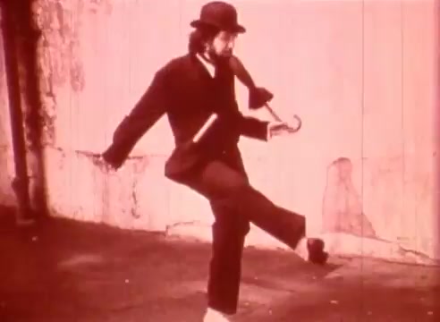 The still depicts a man dressed in vintage attire, including a bowler hat and a coat, playfully walking while balancing a cane. His stance suggests a whimsical and exaggerated movement, typical of silent film comedy. The background is simple and muted, emphasizing the character's expression and movement within the educational context of the film 'Erik Satie and the King of the Beans - Part I'. The overall tone is light and whimsical, reflecting the playful nature of Satie's music.