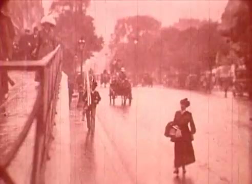 The still from 'Erik Satie and the King of the Beans - Part I' depicts a vintage street scene, likely set in a historic European city. The image shows a rain-soaked promenade with pedestrians, including a woman in period attire holding a bag, and horse-drawn carriages in the background. The color appears faded, contributing to a nostalgic ambiance. The overall atmosphere suggests a leisurely yet damp day, possibly reflecting the everyday life during the time of Erik Satie.