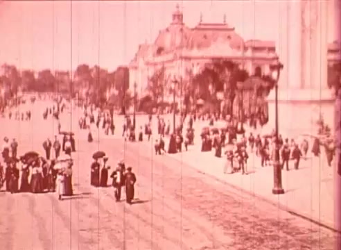 The still from 'Erik Satie and the King of the Beans - Part I' shows a bustling street scene, likely set in a historical period. Large groups of elegantly dressed people, primarily in Victorian-style attire, are seen strolling along a wide avenue. Some individuals carry parasols, indicating a leisurely outing, while notable architecture, possibly a grand building, frames the background. The overall tone is soft and tinted, reminiscent of early 20th-century film techniques, adding a nostalgic quality to the scene.