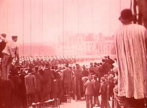 The still from 'Erik Satie and the King of the Beans - Part I' depicts a large crowd assembled in a cityscape, likely for a public event or parade. In the foreground, attendees, dressed in period clothing, watch a formation of marching figures in the background, suggesting a military or ceremonial display. The overall tone is sepia-like, giving the image a historical quality, while the atmosphere is one of anticipation and communal engagement.