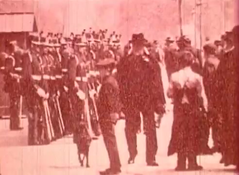 The still from 'Erik Satie and the King of the Beans - Part I' depicts a historical scene featuring a group of uniformed soldiers lined up in formation. In the foreground, a few individuals, possibly leaders or civilians, engage while a small dog is present. The overall tone of the image is sepia-toned, suggesting an older film style typical of educational or documentary formats. The composition reflects a moment of social or military significance, characteristic of the film's exploration of Satie's context and influences.