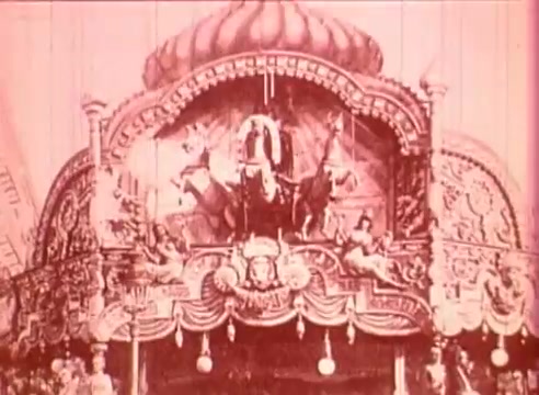 The still from 'Erik Satie and the King of the Beans - Part I' showcases an ornate stage set, likely representing a whimsical or theatrical scene. The design is elaborate, with intricate decorations and figures, suggesting a carnival or festive atmosphere. The color palette has a pinkish hue, enhancing the dreamlike quality of the film. This educational film aims to explore the themes and music of Erik Satie in a visually engaging manner.