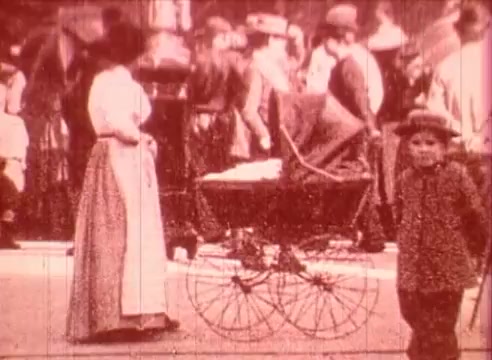 The still from 'Erik Satie and the King of the Beans - Part I' depicts a bustling scene from the early 20th century. In the foreground, a young boy stands near a vintage baby carriage, while a woman in period clothing stands nearby. The background is blurred with a crowd dressed in hats and formal attire, indicative of a lively outdoor event. The color tone suggests an old film quality, enhancing the nostalgic atmosphere of the era.