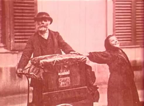 The still from 'Erik Satie and the King of the Beans - Part I' features an elderly man with a beard and a hat, pushing a cart. Next to him is a smiling young girl, joyfully reaching out. The background consists of buildings with shuttered windows, creating a nostalgic and whimsical atmosphere. The overall tone suggests an educational theme, possibly focusing on human connection and the charm of everyday life.