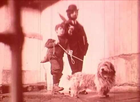 The still from 'Erik Satie and the King of the Beans - Part I' features a sepia-toned scene depicting a boy holding a leash attached to a small dog, while a man in an overcoat and bowler hat stands beside him. The setting appears to be an outdoor area, possibly a street or courtyard, characterized by a simple background. The overall tone of the image evokes a sense of nostalgia and captures a moment of everyday life, typical of early 20th-century films.
