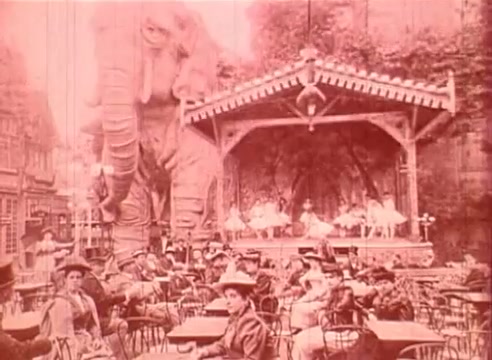The still from 'Erik Satie and the King of the Beans - Part I' showcases a whimsical scene featuring an enormous elephant next to an elevated stage. On the stage, performers in costumes appear to dance or entertain a seated audience scattered at café tables. The setting evokes a nostalgic, circus-like atmosphere, highlighting a blend of nature and entertainment typical of the era. The overall color tone appears pinkish, contributing to the dreamlike quality of the film.