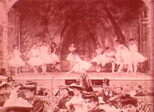 The still from 'Erik Satie and the King of the Beans - Part I' features a lively stage performance, with several dancers dressed in white tutus, showcasing a ballet-like routine. In the foreground, an audience is visible, dressed in period attire, suggesting an atmosphere of entertainment from a bygone era. The overall color tone appears sepia, adding a vintage quality to the scene, which reflects the educational film's emphasis on historical and cultural context.