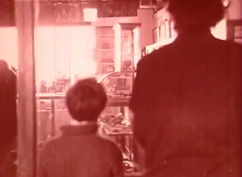 The still from 'Erik Satie and the King of the Beans - Part I' shows a child and an adult walking towards a brightly lit interior, likely a shop or exhibition space. The image has a warm, sepia tone, contributing to a nostalgic atmosphere. Their silhouettes suggest a sense of curiosity and exploration, fitting the educational context of the film. The setting appears filled with objects, hinting at a whimsical or artistic environment related to Satie's imaginative world.