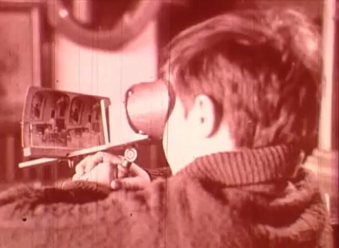The still features a young boy looking intently at a device resembling a projector or viewer. He is holding a tool, possibly for manipulation or adjustment, while seated comfortably. The scene is presented in a warm, reddish hue, suggesting a vintage or nostalgic atmosphere. The educational context implies that the boy is engaging with visual media, likely related to Erik Satie and his artistic contributions, in a learning environment.