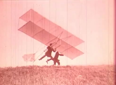The still from 'Erik Satie and the King of the Beans - Part I' shows two figures on a grassy hill, one holding a large glider or flying apparatus. The scene is tinted in a pink hue, evoking a whimsical atmosphere. The figures appear to be in an action pose, suggesting an attempt to launch the glider, reflecting themes of exploration and innovation often associated with early aviation. This educational film likely utilizes such imagery to blend artistic expression with historical context.