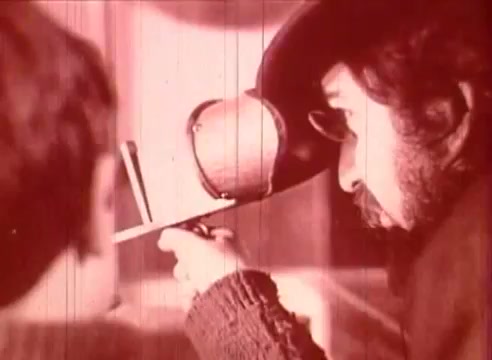 The still from 'Erik Satie and the King of the Beans - Part I' features a man with a beard, wearing a large hat and glasses, intently focused on an object he’s holding. He appears to be demonstrating or examining something, possibly in an educational context, while a younger figure observes him. The image has a reddish tint, typical of certain vintage film styles, and captures a moment of engagement between the two characters.