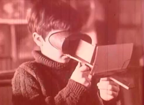 The still from 'Erik Satie and the King of the Beans - Part I' features a young boy wearing a cozy sweater, intently looking through a makeshift viewer made of cardboard. His focused expression suggests curiosity and engagement with the visual experience, emblematic of the educational theme of the film. The warm-toned color palette enhances the nostalgic feel of the scene.