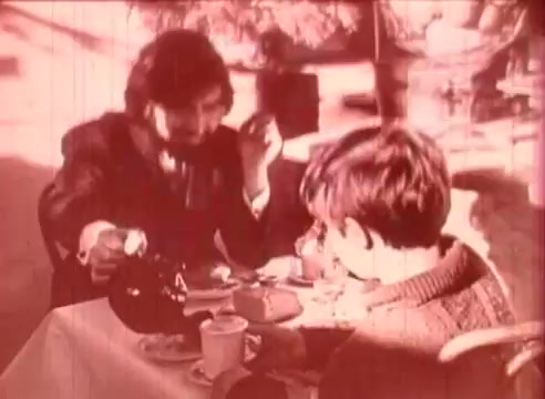 In this 16mm still from 'Erik Satie and the King of the Beans - Part I,' a man, possibly portraying Erik Satie, is animatedly engaging with a young boy at a table set with cups and saucers. The scene suggests a warm and educational interaction over tea, infused with a playful atmosphere. The visual style is characterized by a soft, nostalgic hue, indicative of the film's vintage aesthetic.