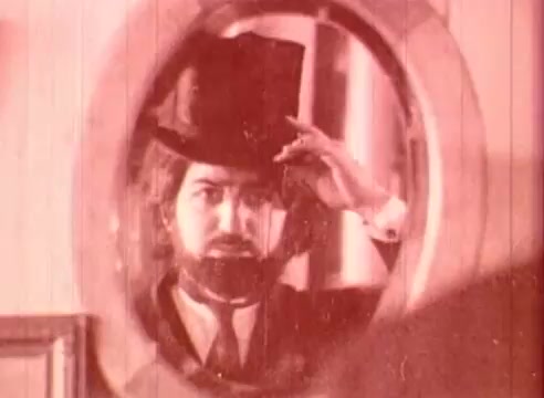 The still from 'Erik Satie and the King of the Beans - Part I' features a man with a beard and long hair, dressed in formal attire, including a top hat. He is looking at his reflection in a circular mirror, adjusting his hat. The image has a sepia-toned or red-tinted quality, contributing to a vintage aesthetic. This scene likely highlights a moment of self-reflection or preparation, aligning with the film's educational themes centered around the composer Erik Satie.