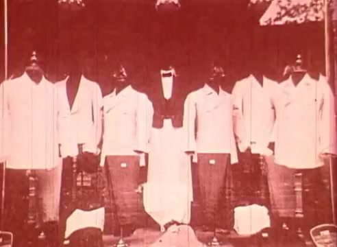 The still from 'Erik Satie and the King of the Beans - Part I' features a series of figures dressed in formal attire, likely representing a group. The image appears to be in a faded color palette, adding a vintage feel. The context suggests an educational focus on Satie's life or artistic influences, perhaps using visual storytelling to illustrate themes related to his work or era. The symmetrical arrangement of the figures may symbolize order or tradition in contrast to Satie's innovative spirit.