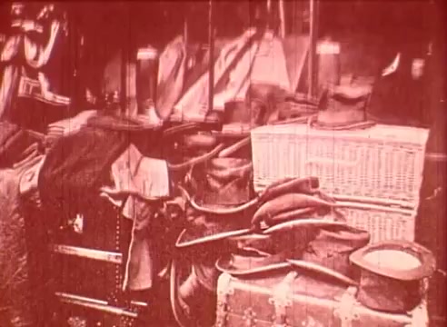The still from 'Erik Satie and the King of the Beans - Part I' features a cluttered assortment of various objects. The image is monochromatic with a reddish tint, showcasing items like hats, bags, and crates. This scene likely reflects a whimsical or chaotic environment, aligning with the film's educational and artistic themes related to Erik Satie. The overall composition conveys a sense of playful disarray, inviting curiosity about the story being told.