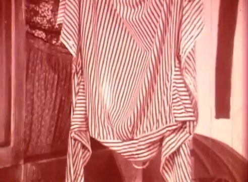 The still features a striped garment hanging prominently in a room, surrounded by wooden furniture and additional fabric. The reddish tint suggests a vintage aesthetic, characteristic of 16mm film. This scene likely serves to evoke a whimsical or playful atmosphere, aligning with the educational goals of 'Erik Satie and the King of the Beans - Part I.'