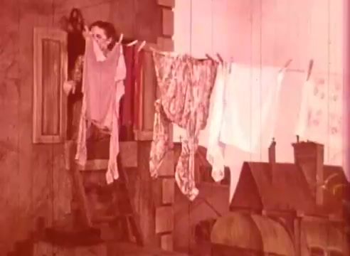 The still from 'Erik Satie and the King of the Beans - Part I' features a character standing on a ladder, seemingly peering out from a window. Clothes hang on a line nearby, adding a domestic scene. The warm, reddish tint gives it a vintage feel, indicative of its educational and artistic context. The setting appears whimsical, reflecting elements of storytelling associated with Erik Satie's musical style.