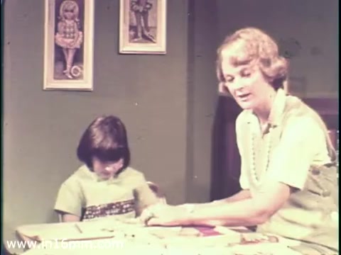 The still depicts a woman interacting with a child at a table, likely engaged in an educational or creative activity. The woman appears to be explaining something, while the child, seemingly focused or somber, listens attentively. The setting includes simple decorations, like framed images on the wall, reflecting a domestic environment. This scene is likely part of a larger public service announcement aimed at raising awareness about measles and its potential impact on mental disability.
