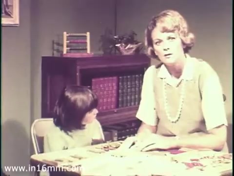 The still from the 1968 PSA features a concerned woman sitting at a table with a young girl. The setting appears to be a home, and the backdrop includes shelves with books and educational materials. The woman is likely discussing the dangers of measles and its potential impact on mental disabilities, capturing a serious and informative tone typical of public service announcements from that era. The image conveys a sense of urgency regarding children's health and education.