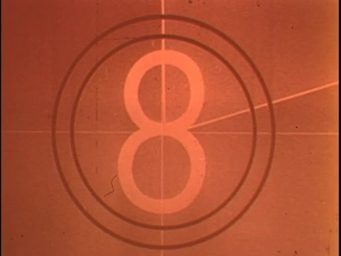 The still appears to be a countdown leader, featuring the number '8' in a circular format, indicative of old film stock. The image has a warm, reddish tint likely due to aging or the negative format. The design includes crosshairs, typical of countdown leaders used in film production, serving as a reference for syncing sound and vision. This type of element is essential for preparing viewers for the start of a commercial or film sequence. For the Koogle Flavored Peanut Butter commercial, consider enhancing the contrast and adjusting color balance to achieve a more neutral look, while ensuring the countdown remains visible against the tinted background.