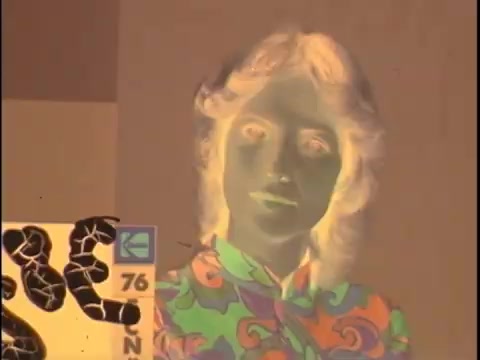 In the image from the Koogle Flavored Peanut Butter commercial negative, the colors are inverted due to the negative film process. The central figure is a person with light-colored hair styled in soft curls, wearing a colorful, psychedelic-patterned shirt, prominently featuring vibrant swirls and shapes.   On the left, there's a black-and-white visual element, possibly related to branding or a graphic design, with abstract shapes that may symbolize the product. The features of the person and any surrounding details appear surreal and stylized due to the color inversion, enhancing the playful, nostalgic feel of the commercial.   For your editing project, focus on restoring the original colors to reflect the whimsical and bold aesthetic typical of advertisements from that era, while syncing with the sound for
