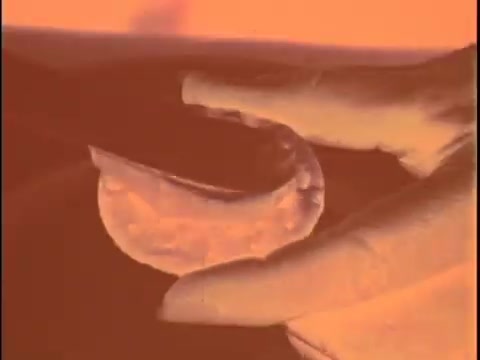 This still from the 'Koogle Flavored Peanut Butter' commercial features a close-up of a hand holding a circular peanut butter product, likely showcasing its creamy texture. The image has a vintage feel due to the 16mm film format, and the color tone appears to lean towards orange, which may indicate it’s a negative or an uncorrected image. The focus seems to be on the product itself, emphasizing its unique features.   For color correction, consider enhancing the contrast and adjusting the color balance to bring out the original colors, particularly the peanut butter's warm tones. Focus on removing the orange cast and restoring a more natural appearance.