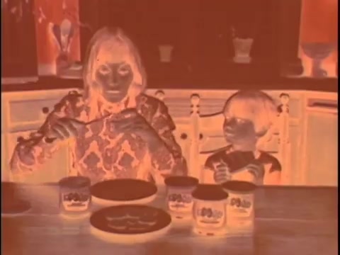 The still from the 'Koogle Flavored Peanut Butter' commercial presents a negative image of a scene featuring a woman and a boy at a kitchen table. They appear to be engaged with jars of Koogle peanut butter. The woman, in a patterned outfit, is holding a spoon, while the boy is looking on with an expression of curiosity. The jars are prominently displayed on the table, hinting at the product's promotional focus. The kitchen setting is identifiable, characterized by cabinetry and some background decor, although the colors are inverted due to the negative format, giving the scene an unusual and surreal appearance.   For color correction, consider reintroducing warm tones to reflect the typical peanut butter hues, along with adjusting skin tones for realism. Ensure that