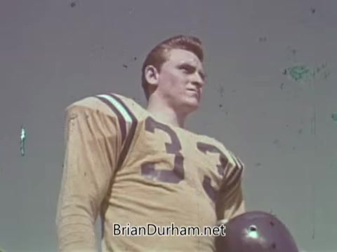 The still captures a muscular, athletic figure dressed in a vintage football uniform, featuring a yellow jersey with purple stripes and the number '33.' The character, possibly meant to symbolize strength or safety in automotive contexts, gazes intently off-screen against a clear blue sky. The overall tone suggests a focus on safety and preparedness, typical of public service announcements from that era.
