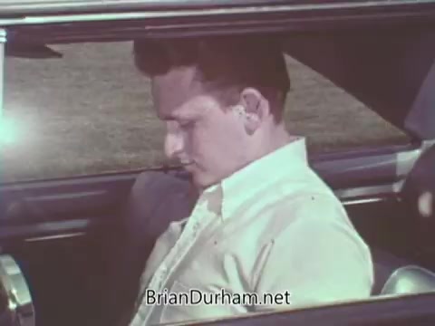 The still from the 'Automotive Safety Foundation PSA' depicts a young man sitting in a car, appearing contemplative or slightly pensive. He is dressed in a light-colored shirt, and the interior of the car is visible, showcasing classic design elements. The background suggests an outdoor setting with grass, hinting at a serene environment. This PSA likely emphasizes safety awareness and responsible driving behaviors common in public service announcements from the 1960s.