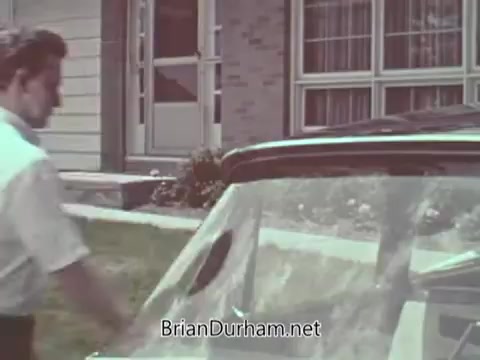 The still captures a man inspecting the windshield of a car parked in a residential area, emphasizing automotive safety. The background features a suburban house, indicating a domestic setting. The focus on the windshield suggests a theme of vehicle safety awareness, typical of public service announcements from the 1960s.