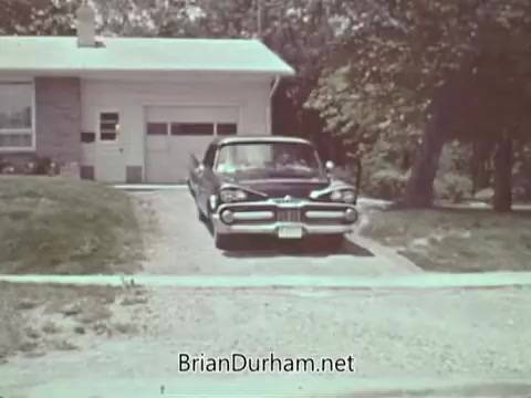 The still from the 1960s PSA by the Automotive Safety Foundation features a classic car parked in a driveway alongside a suburban home. The scene evokes a mid-20th-century American residential setting, highlighting the emphasis on automotive safety during that era. The greenery and the driveway suggest a focus on neighborhood safety and responsible driving practices.