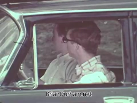 The still shows two men seated in the front of a car, highlighting an automotive safety message typical of a 1960s public service announcement. The scene emphasizes the importance of safety in vehicles, likely portraying a discussion about driving practices or safety features. The men’s attire reflects the fashion of that era, enhancing the nostalgic context of the PSA.