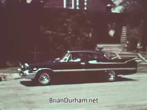 The still features a classic 1960s car parked on a residential street. The vehicle stands out with its vintage design, showcasing chrome details and a bold color scheme, typical of that era. The background includes a small house and greenery, creating an everyday suburban scene. The overall mood reflects a focus on automotive culture during that time, likely emphasizing safety in driving.