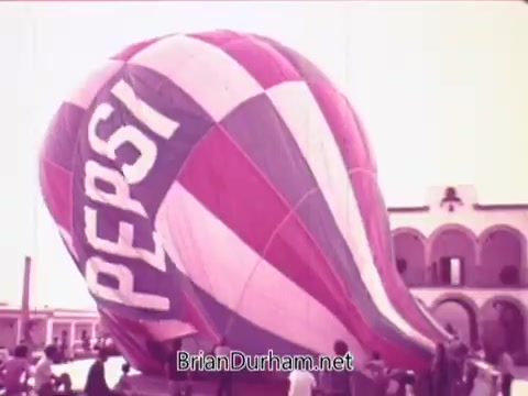 The still features a large hot air balloon adorned with the Pepsi logo. The balloon is vividly colored in pink, purple, and blue stripes and appears to be in the process of being inflated. A group of people gathers around the balloon, likely participating in or observing an event. The setting seems festive, aligning with the theme of the commercial titled 'Todos junto a Pepsi siempre libre y feliz' (Everyone with Pepsi always free and happy).