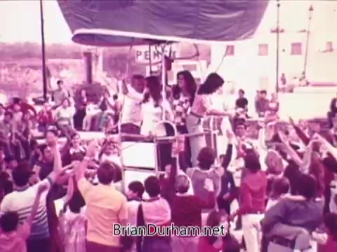 The still from the Pepsi commercial features a lively outdoor scene with a crowd enthusiastically reaching out towards a small group of people on a float or platform. The individuals on the float appear to be engaging with the crowd, creating a festive atmosphere. The setting suggests a celebration, with many participants dressed in colorful clothing, reinforcing a sense of community and joy associated with the brand.