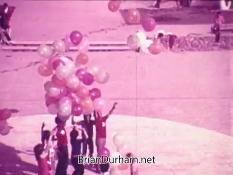 The still shows a vibrant outdoor scene featuring a group of people wearing matching red shirts, joyfully reaching up to a cluster of colorful balloons. The image has a soft, pinkish tone, creating a cheerful and festive atmosphere. In the background, a few individuals are scattered, contributing to a lively, communal spirit associated with the promotional theme of Pepsi.