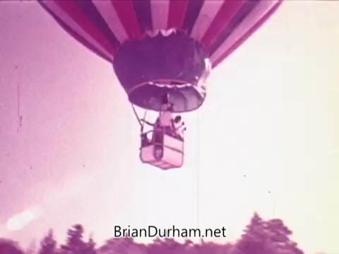 The still from the 'Pepsi commercial - Todos junto a Pepsi siempre libre y feliz' features a colorful hot air balloon in flight. The balloon is adorned with vibrant stripes, and a small basket can be seen below it, likely carrying a person. The background is filled with a soft, pastel sky, suggesting a serene and uplifting atmosphere. This scene conveys a sense of freedom and joy, aligning with the themes of the commercial.